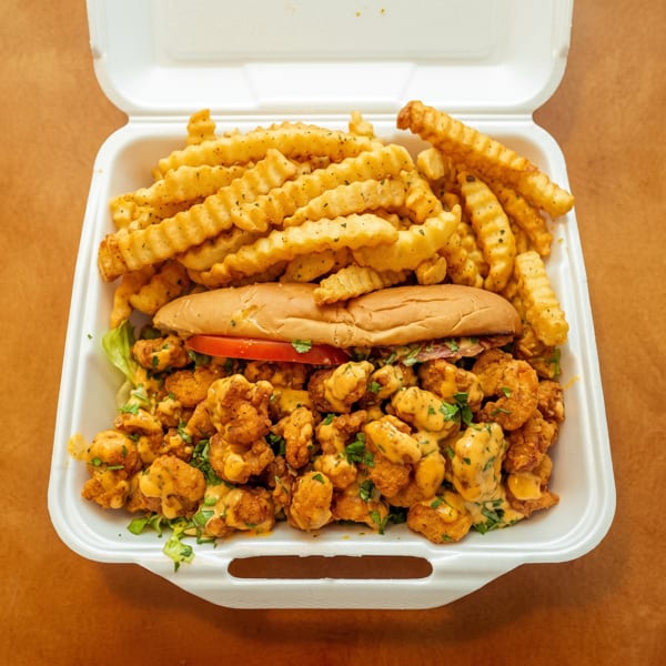 Takeout container with a shrimp po'boy, sauced fried shrimp, and seasoned crinkle-cut fries.