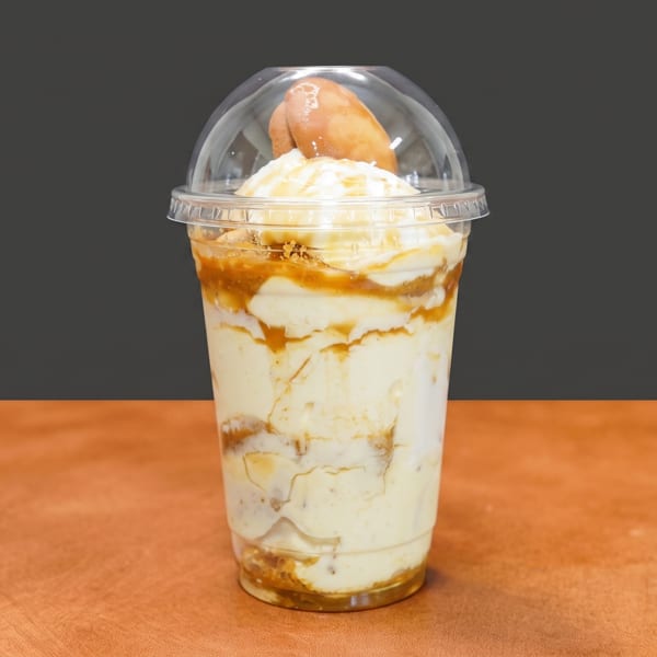 Creamy layered dessert in a plastic cup with caramel swirls, topped with a pastry and clear dome lid.