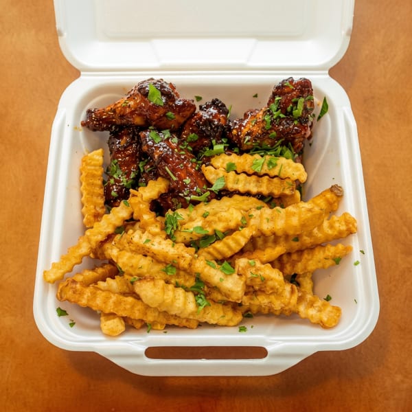 Glazed chicken wings and crinkle-cut fries with herbs in a white takeout container.