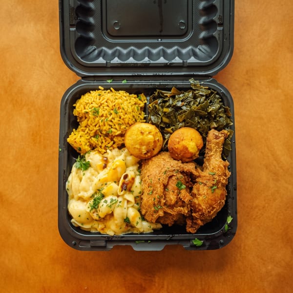 Southern comfort meal with fried chicken, mac and cheese, rice, collard greens, and hush puppies in a takeout container.
