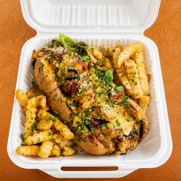 A cheesy, loaded sub sandwich with crinkle-cut fries, garnished with herbs, in a white takeout container.