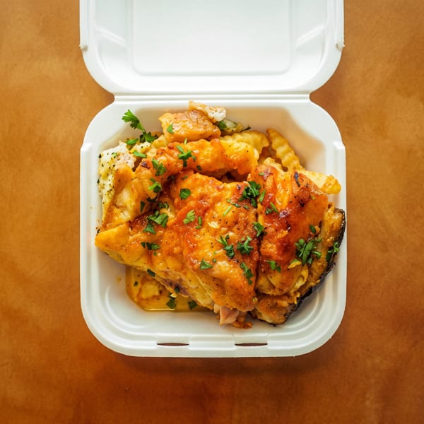 Open white takeout container with sauced chicken, crinkle-cut fries, and parsley on a brown surface.
